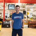 A man wearing the America est. 1776 shirt stands and smiles inside a casual office space.