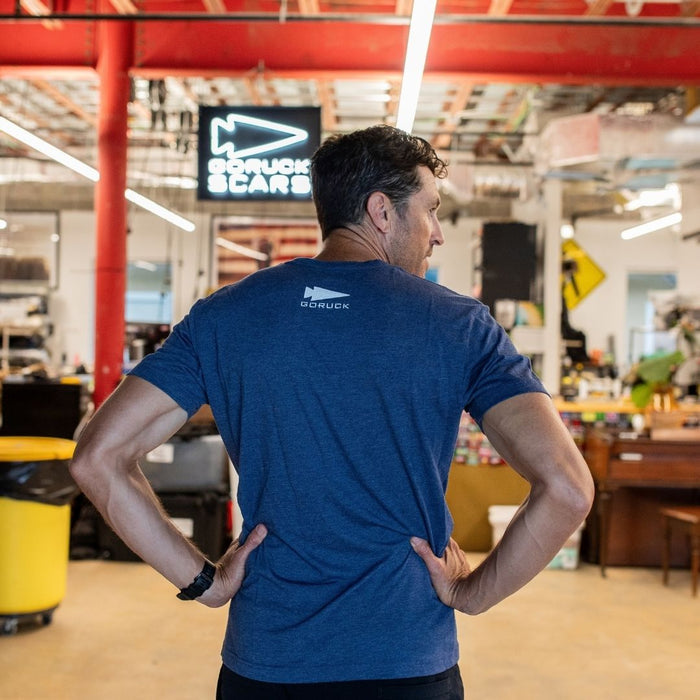 A man in a blue GORUCK Spearhead Tee - Stars & Stripes - Tri-BlendX stands with his hands on his hips inside a workshop filled with red beams and tools, the shirt’s spearhead emblem subtly honoring his Special Forces background.