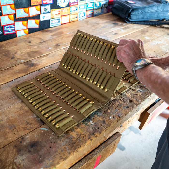 A person opens an Ammo Book filled with rifle ammunition on a wooden table in a workshop decorated with patches on the wall.