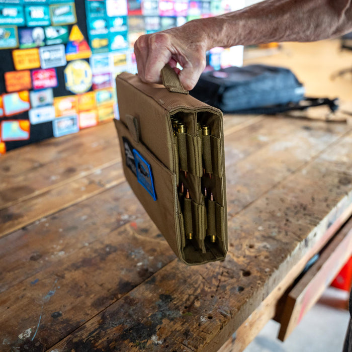 A hand lifts the Ammo Book, a tan book-style organizer with brass objects inside, from a wooden workbench with patches in the background.