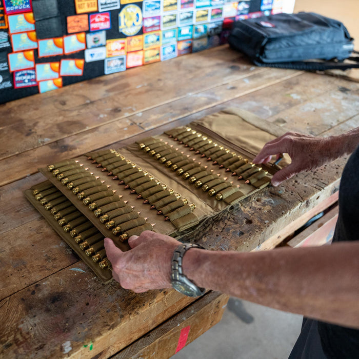 A person arranges shotgun shells in an Ammo Book on a wooden workbench decorated with travel stickers.