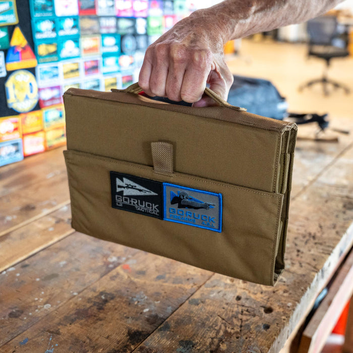 A hand holds a brown Ammo Book organizer with patches on a wooden table in a colorful workspace.