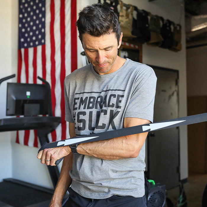 Man in gym wearing "Embrace the Suck" shirt, stretching with a Recovery Floss Band to improve mobility, with a U.S. flag in the background.