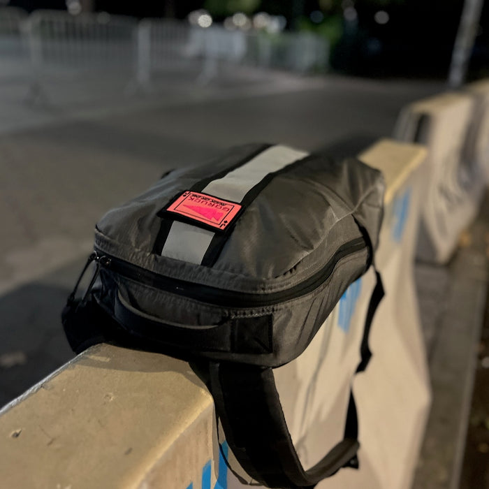 A Basic Rucker in gray, featuring a reflective stripe and a pink label, rests on a concrete barrier at night—ready for your next GORUCK rucking adventure.