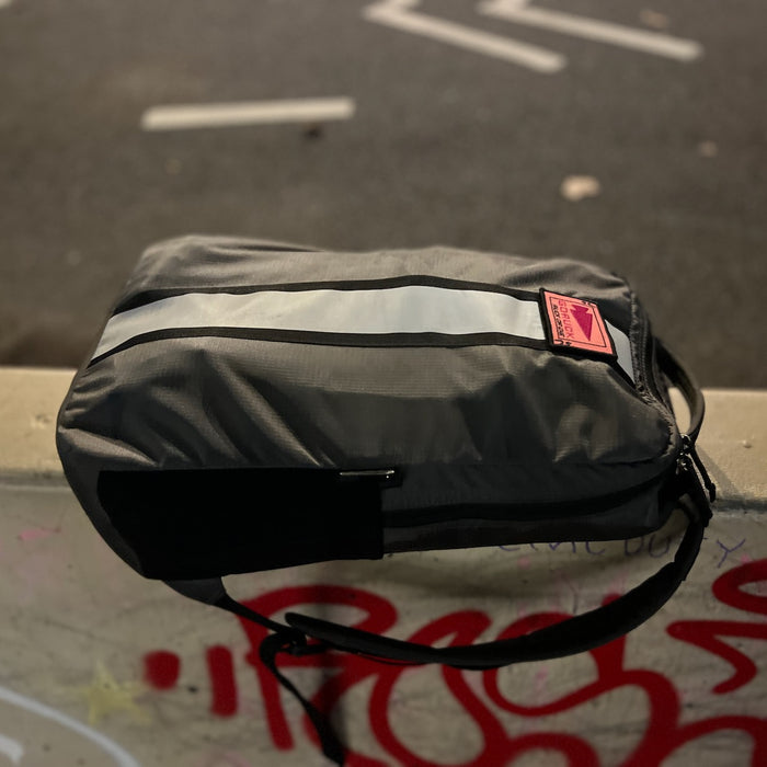 A black and gray Basic Rucker by GORUCK, featuring a reflective stripe, rests on a graffiti-covered concrete ledge at night—ideal for rucking or everyday use.