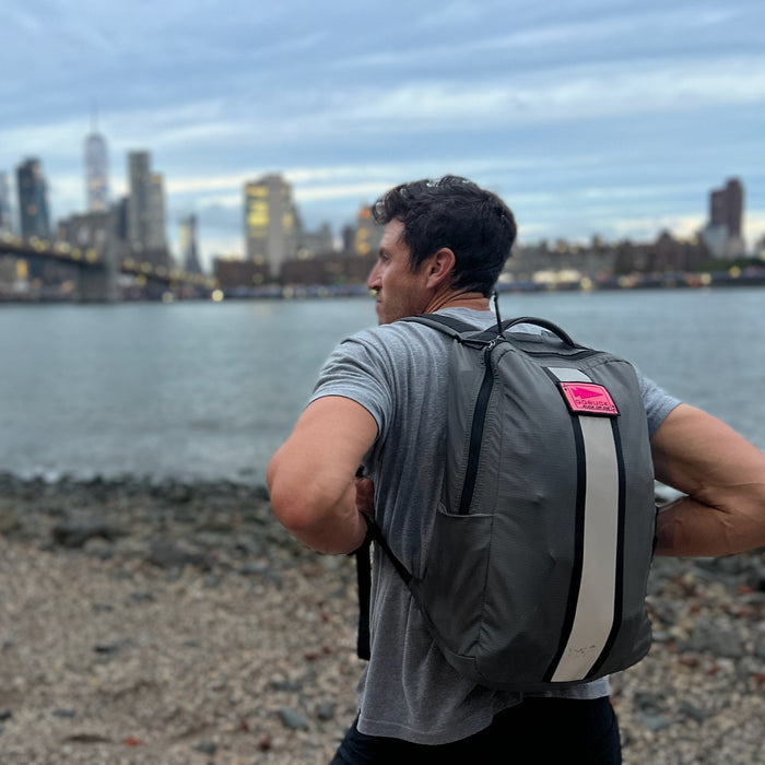 A man wearing a gray Basic Rucker backpack stands on a rocky shore with a city skyline and bridge in the background, embodying the spirit of rucking amidst urban adventure.