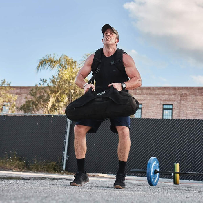 Man lifting GORUCK sandbag outdoors in black ballistic trainers, fitness gear focus