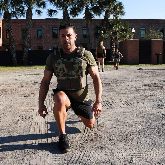 Man in GORUCK weighted vest performing outdoor lunge exercise on dirt, palm trees in background
