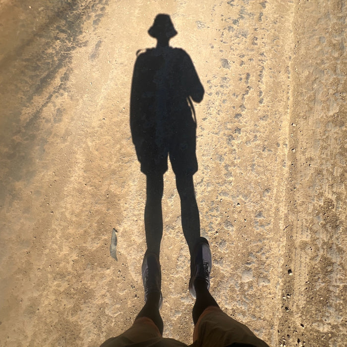 Long shadow of a person wearing the Boonie Hat - Slick - ToughDry on a sunlit dirt road, their feet visible at the bottom of the image.