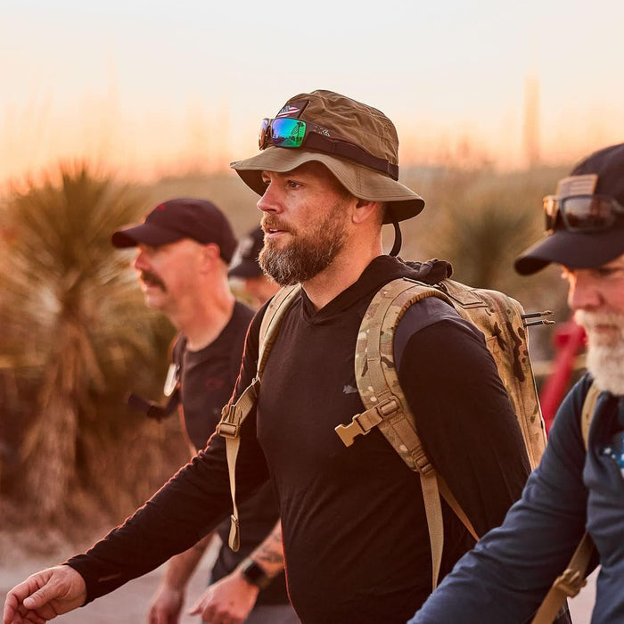 Man wearing ranger green boonie hat, black shirt, and camouflage backpack, rucking outdoors at sunset, GORUCK gear in use.
