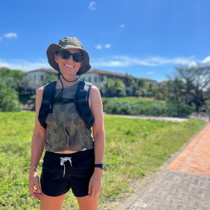 A smiling person wearing the Boonie Hat - Tactical - ToughDry, sunglasses, and a backpack stands outdoors on a sunny day, surrounded by lush greenery.