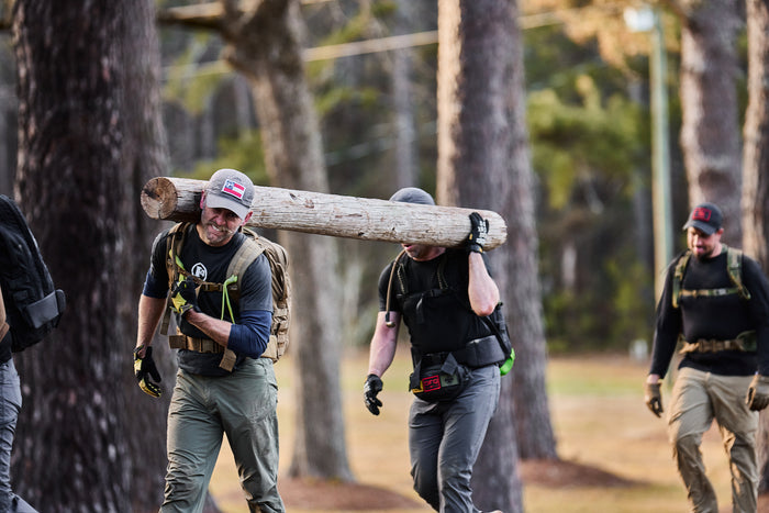 Three men in outdoor gear carry a large log together through a wooded area during a BRAGG 2027 team exercise inspired by Special Forces training.