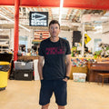 A man wearing a black t-shirt and shorts stands in an industrial workspace with a Buddy FCKR Friday sign behind him.