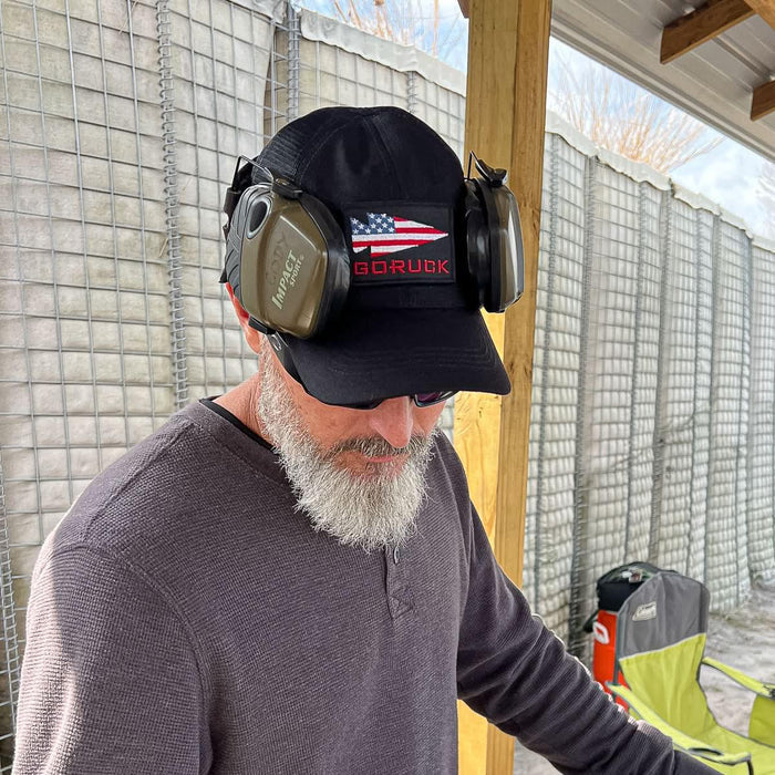 Man wearing GORUCK black hat with American flag patch, earmuffs, and sunglasses outdoors