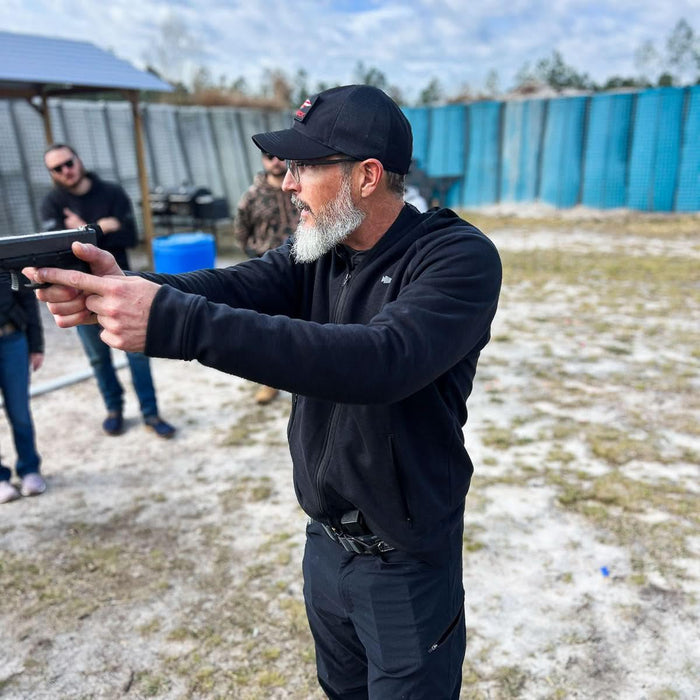 Man in black GORUCK hat and jacket aiming a pistol outdoors at training event