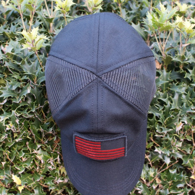 A Cadre Hat - CORDURA®, featuring a black military-grade design and a red patch, rests atop green, spiky holly leaves.