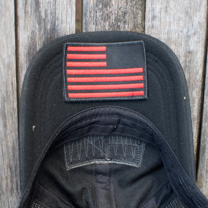 The Cadre Hat - CORDURA® is a black, military-grade cap featuring a red, stylized American flag patch. It’s crafted from rugged 500D CORDURA® fabric and shown on a weathered wooden surface.