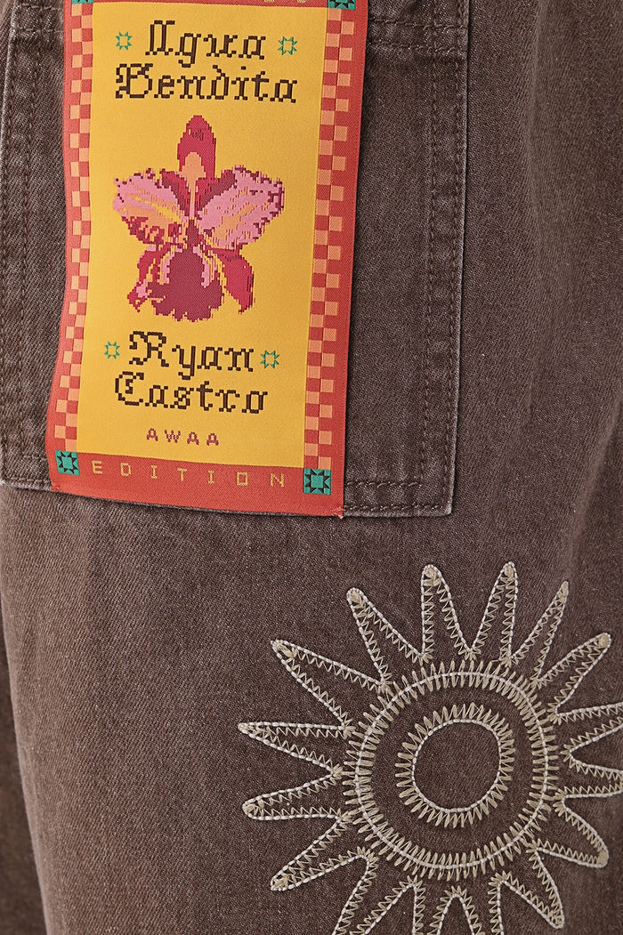Brown men's shorts with white embroidered sun design on front and woven label featuring pink orchid on checkered border