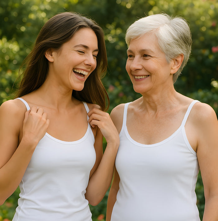 Two women, one younger and one older with short gray hair, smile together outdoors in Teri Lingerie Cozy Cotton Camisole (Style 1500) white tops, surrounded by lush greenery.