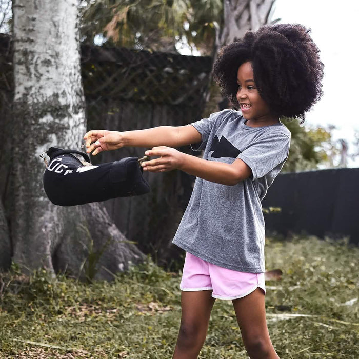 Smiling child using GORUCK training sandbag outdoors, wearing gray t-shirt and pink shorts