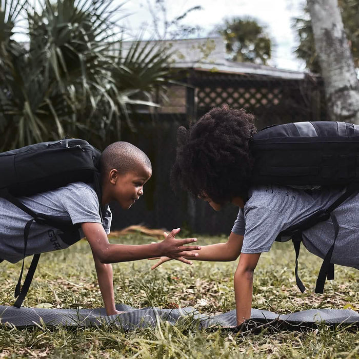 Two kids wearing black GORUCK backpacks do push-ups and high-five outdoors on grass.
