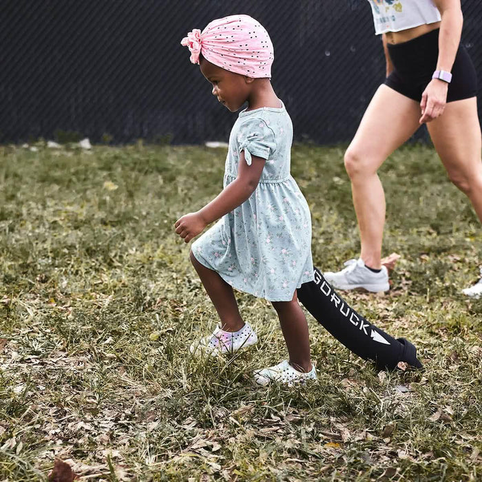 Child in floral dress and pink headwrap outdoors, walking near GORUCK rucking sandbag