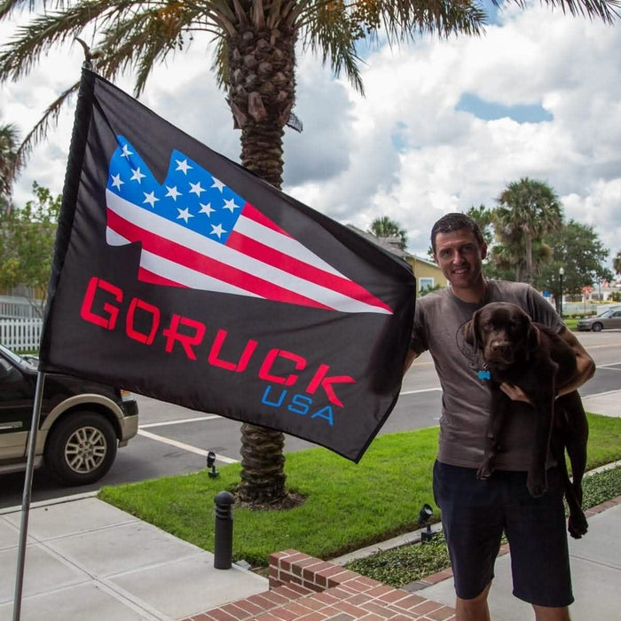 Man holding GORUCK USA flag and a brown dog outdoors near palm trees and street