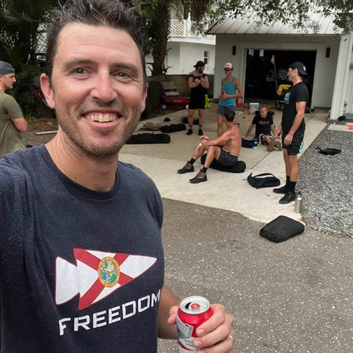 Man in GORUCK Freedom shirt smiling, holding a drink, with group rucking outdoors near garage.
