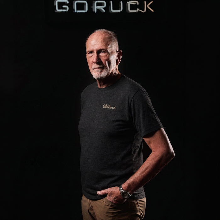 An older man wears the GORUCK Bud Tee - Tri-BlendX, standing against a black background with a "GORUCK" sign above him.
