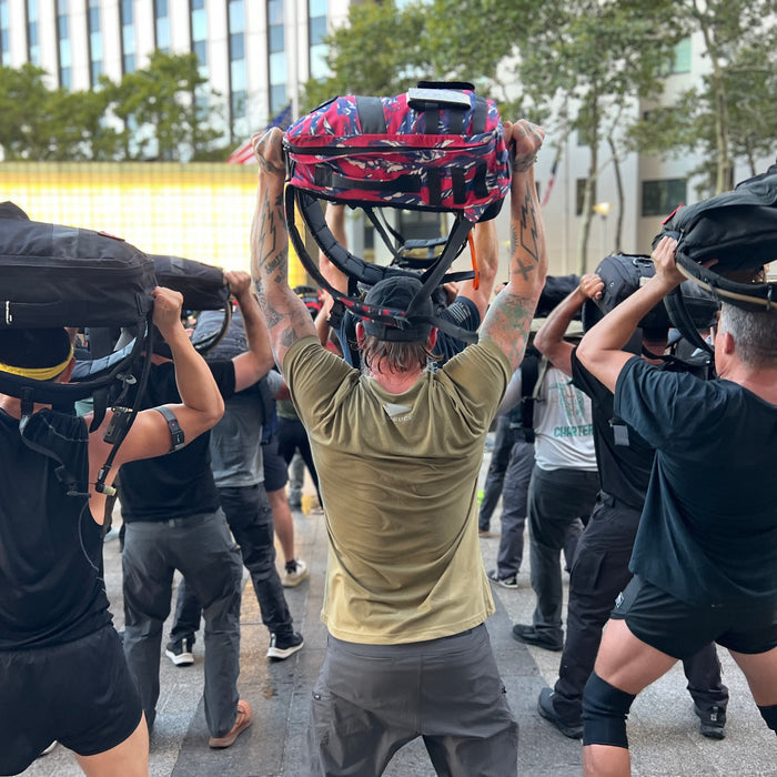 A group of people outdoors, facing away from the camera, lifts backpacks and helmets overhead—several are wearing hi-vis GORUCK Reflective Tee - Tri-Blend shirts reminiscent of Special Forces teams.