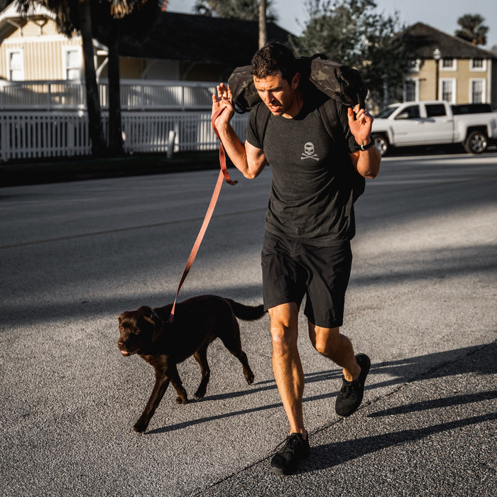 Wearing the GORUCK Tribe Tee - Tri-BlendX, a man carries weight on his shoulders as he walks a brown dog on a leash down a sunny street, embodying the spirit of the GORUCK Tribe and community.
