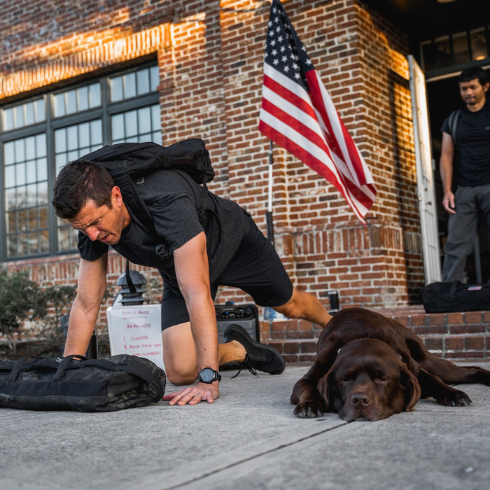 Wearing the GORUCK Tribe Tee - Tri-BlendX, a man does push-ups on the sidewalk next to a resting brown dog, with the U.S. flag and another man in the background—highlighting the true sense of community at every GORUCK Challenge.