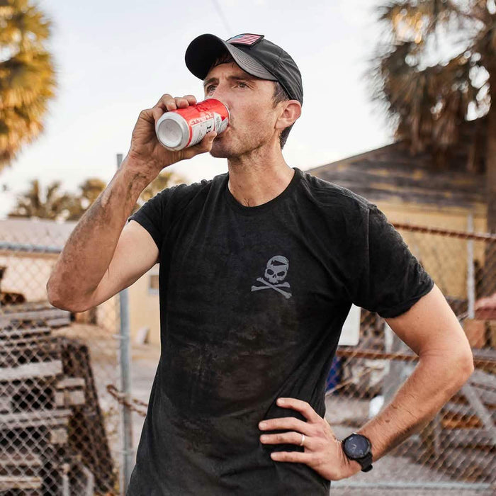 Man wearing GORUCK black skull logo tee outdoors, drinking from a can, with American flag cap.