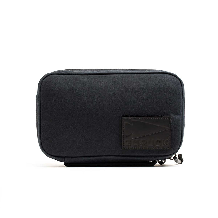 The GR1 Field Pocket - Waxed Canvas is a black, rectangular pouch made of waxed canvas, with a stitched logo patch on the front and a zipper along the bottom edge.