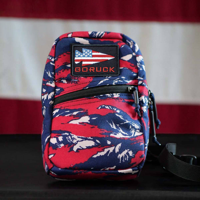 A GRenade Ruck - American Tiger Stripe, small and featuring an American flag patch, sits in front of a US flag background.