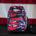 A GRenade Ruck - American Tiger Stripe, small and featuring an American flag patch, sits in front of a US flag background.