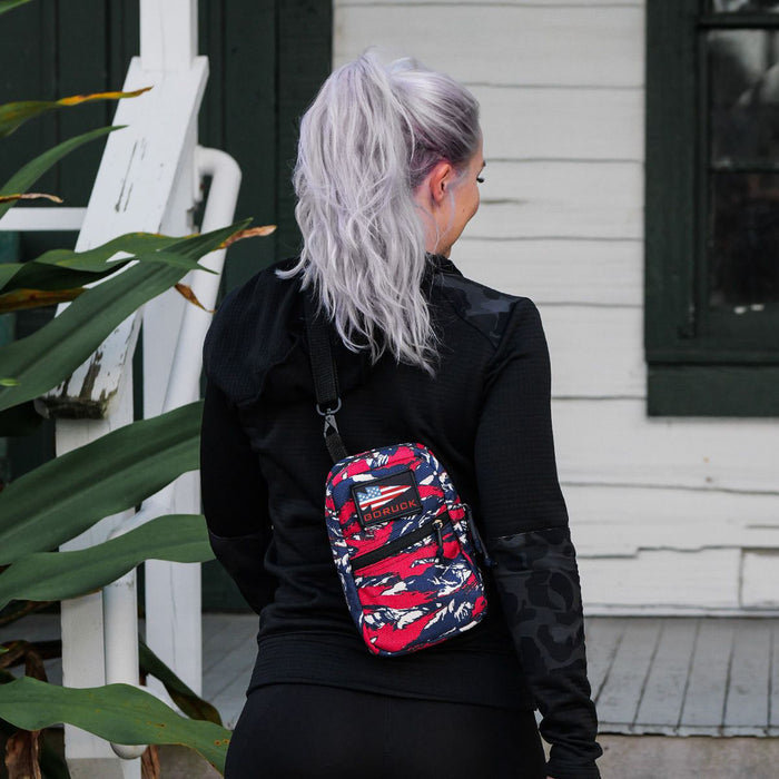 A woman with long, silver hair stands outside a white house, wearing the GRenade Ruck - American Tiger Stripe cross-body bag.