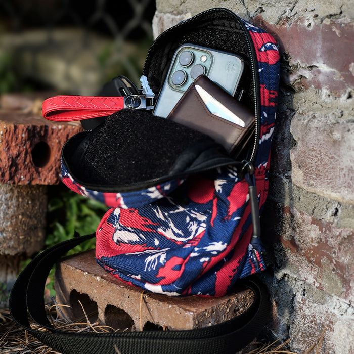 A GRenade Ruck - American Tiger Stripe sits on a brick next to a wall, holding a phone, wallet, and red multitool.
