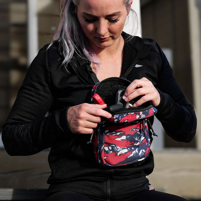 Woman in a black jacket places her phone into an American Tiger Stripe GRenade Ruck cross-body bag while sitting outside.