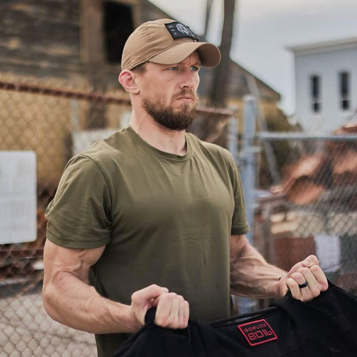 Man outdoors wearing a GORUCK hat and olive shirt, lifting rucking gear, rugged urban background