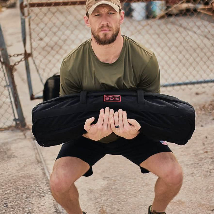 Man outdoors lifting GORUCK ruck sandbag, fitness training gear, squatting by chain-link fence