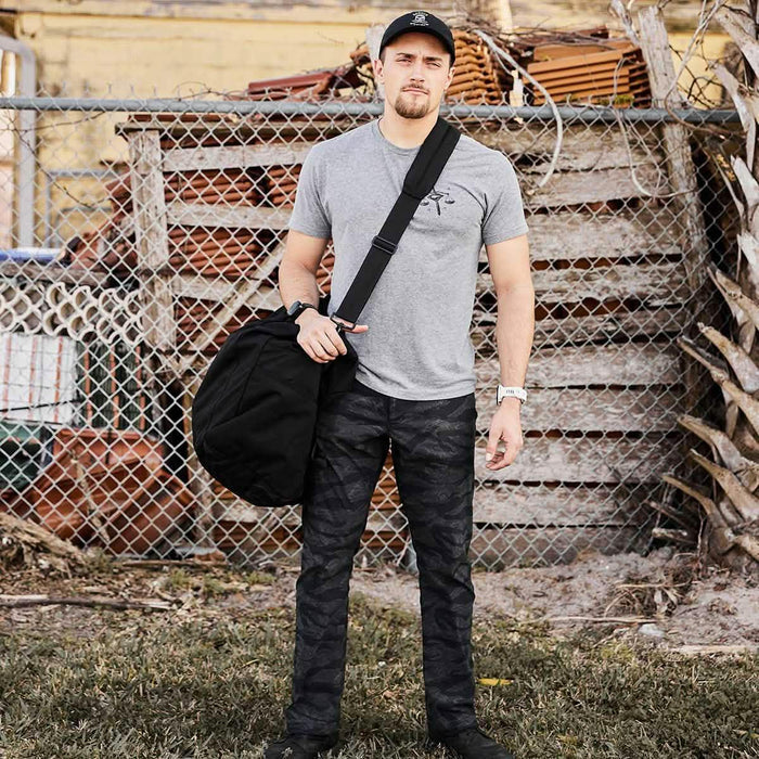 Man in GORUCK gear with rucksack standing outdoors by fence, wearing gray shirt and camo pants
