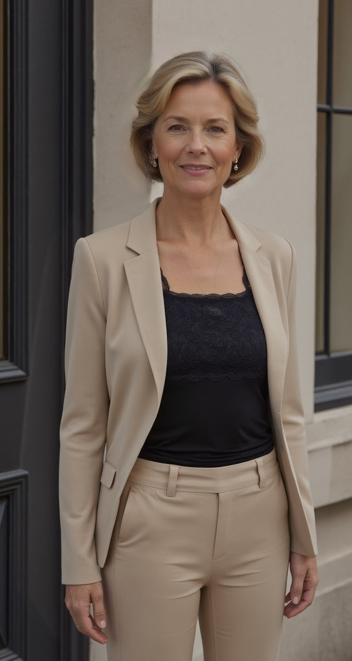 A woman with short blonde hair stands outside against a building wall, smiling slightly at the camera. She wears a beige suit and the Lace Layering Camisole (Style 1506) by Teri Lingerie.