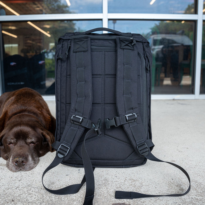 The M26 PREORDER // Q4 2026 ETA, a black backpack with a bombproof laptop compartment, stands upright on concrete beside a sleeping brown dog in front of glass doors.