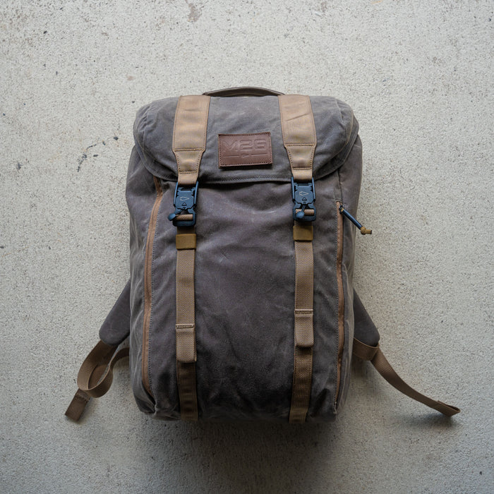 The M26 PREORDER // Q4 2026 ETA is a brown canvas backpack with tan straps and a leather patch, shown on a light concrete surface and featuring a bombproof laptop compartment for extra protection.