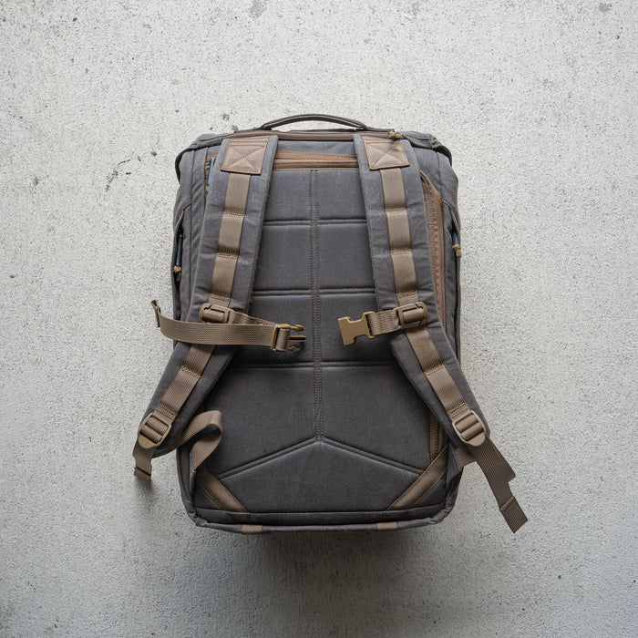 The M26 PREORDER // Q4 2026 ETA backpack, featuring padded straps, buckles, and a bombproof laptop compartment, is displayed flat on a textured light-colored surface.