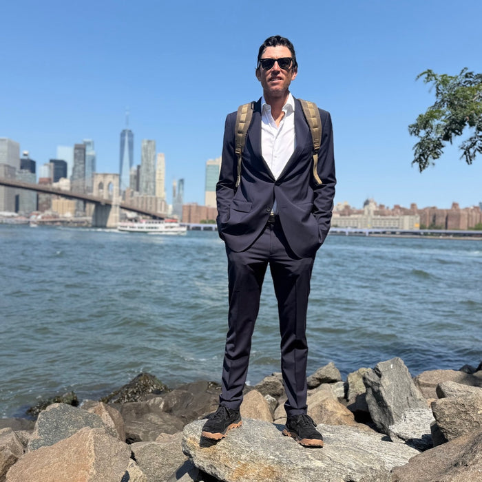 A man wearing a suit and backpack, sporting the Mackall 45 Black + Gum shoes, stands on rocks by a river with a city skyline and bridge in the background under a blue sky.