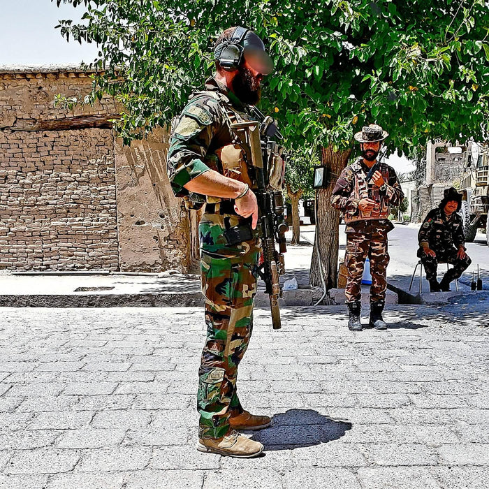 Men in tactical gear and camouflage stand outdoors by brick building and trees, demonstrating GORUCK rugged rucking equipment.