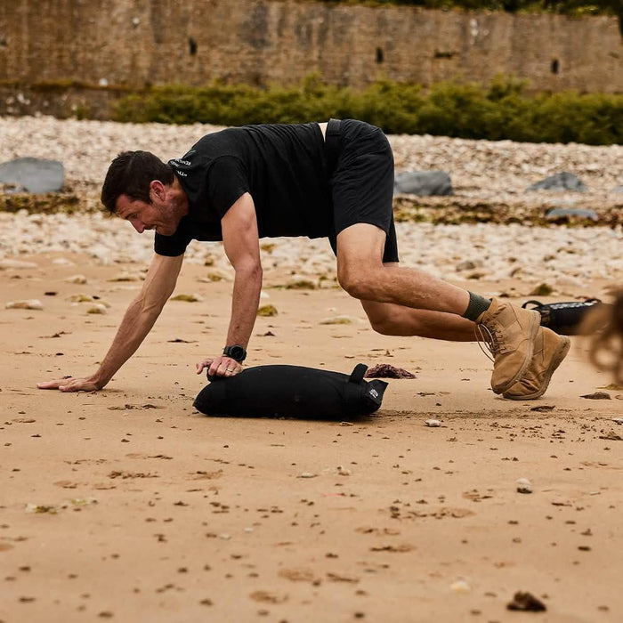 Man training on beach with GORUCK sandbag, wearing black gear and rugged boots