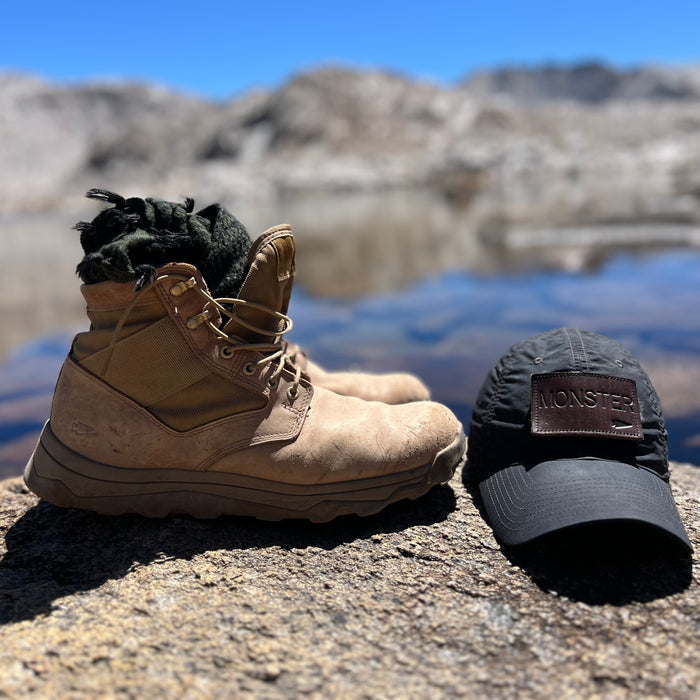 The MACV-2 Mid Top boots in Briar + Coyote and a black “MONSTER” cap sit atop a lakeside rock with mountains in the background, evoking the rugged design of classic Special Forces footwear such as the legendary Vietnam jungle boot.